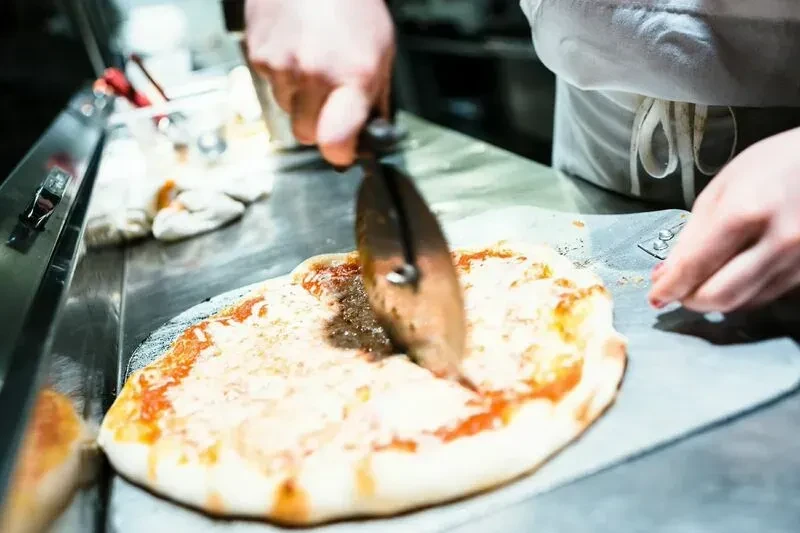 Hands slicing a freshly baked, cheesy pizza with a cutter.