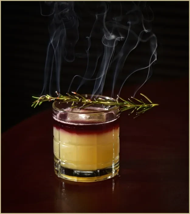 Three-layered cocktail in a faceted glass, garnished with smoking rosemary.