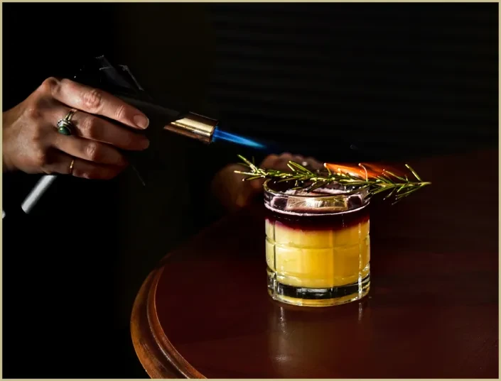 Layered cocktail with rosemary and orange peel is being flamed.