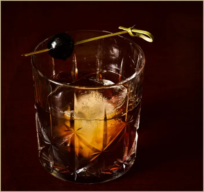 Dark amber cocktail in crystal glass with large ice and dark garnish.