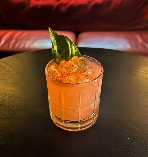 Orange cocktail in faceted glass garnished with basil and spice.