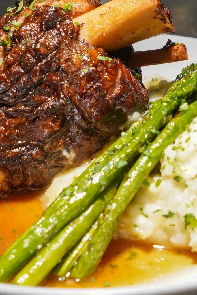 Roasted bone-in meat, mashed potatoes, asparagus, and sauce, garnished with herbs.
