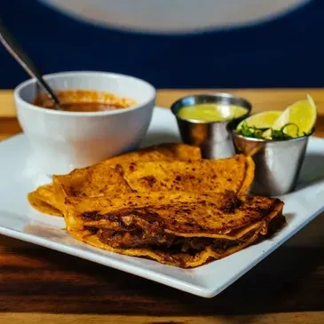 Two crispy birria tacos with consommé, lime, cilantro, and green sauce.