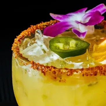Close-up: yellow-green cocktail with spiced rim, jalapeño, and purple orchid.