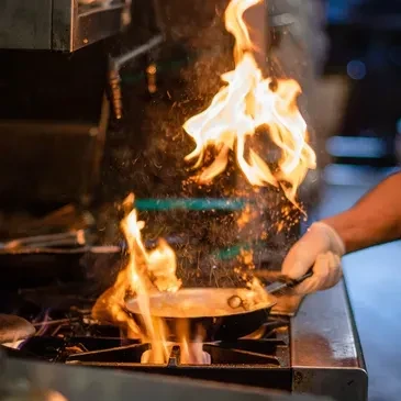 Chef's hand flambéing food in a pan over a gas burner.