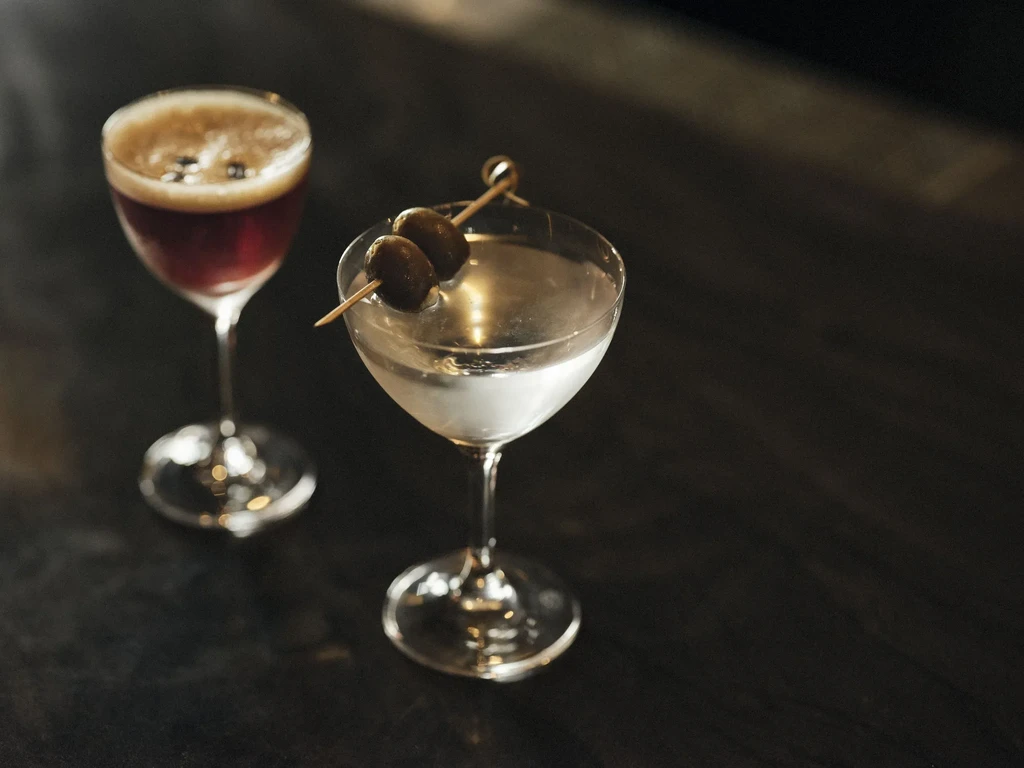Two cocktails in coupe glasses, one with olives, one with foam.