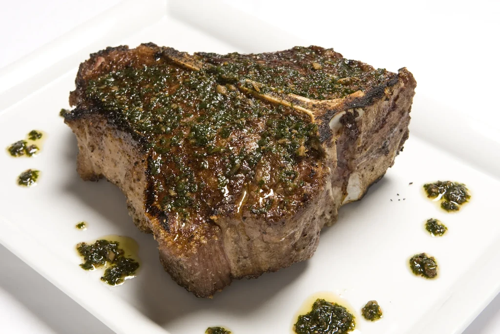 Thick-cut grilled T-bone steak with green herb sauce on a white plate.