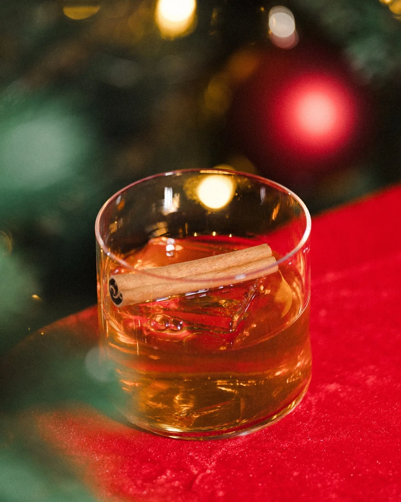 Amber beverage with ice, cinnamon stick, on red surface, festive setting.