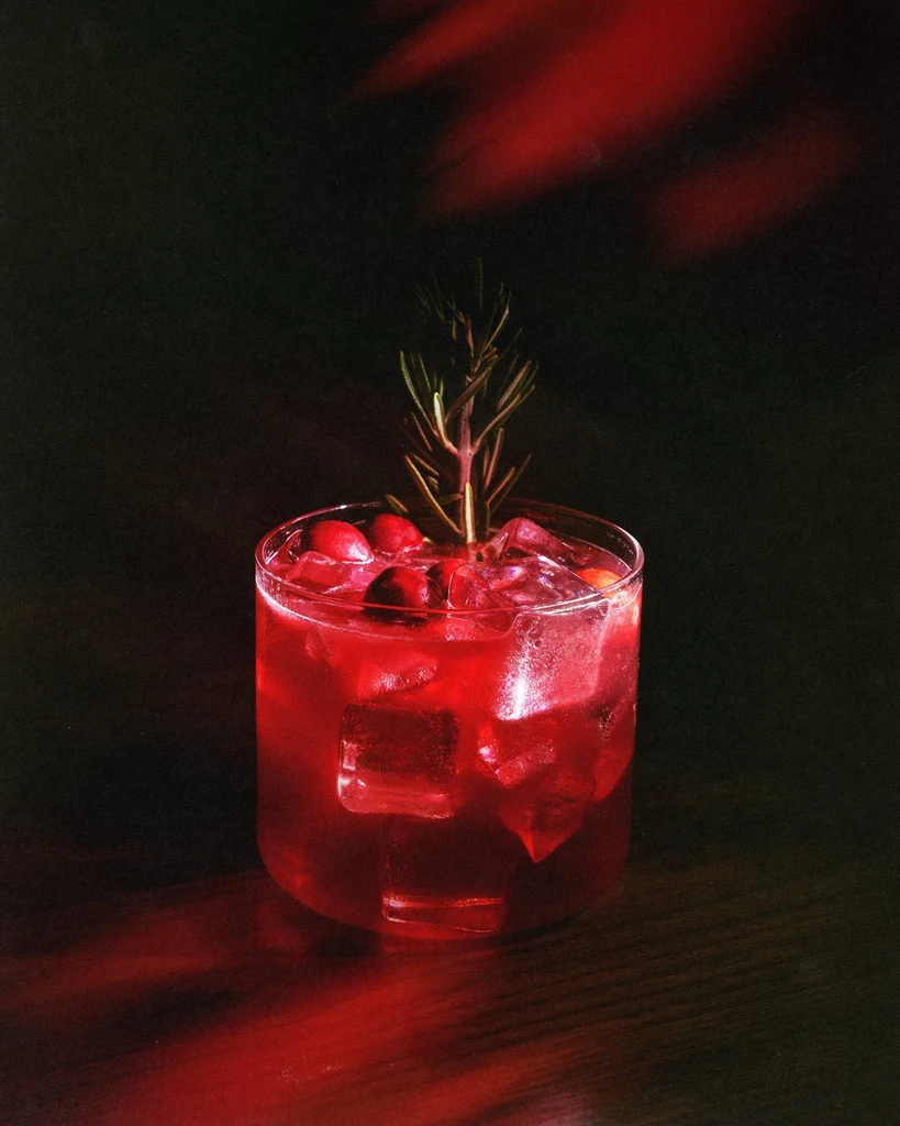 Red cocktail with ice, berries, and rosemary in a rocks glass.