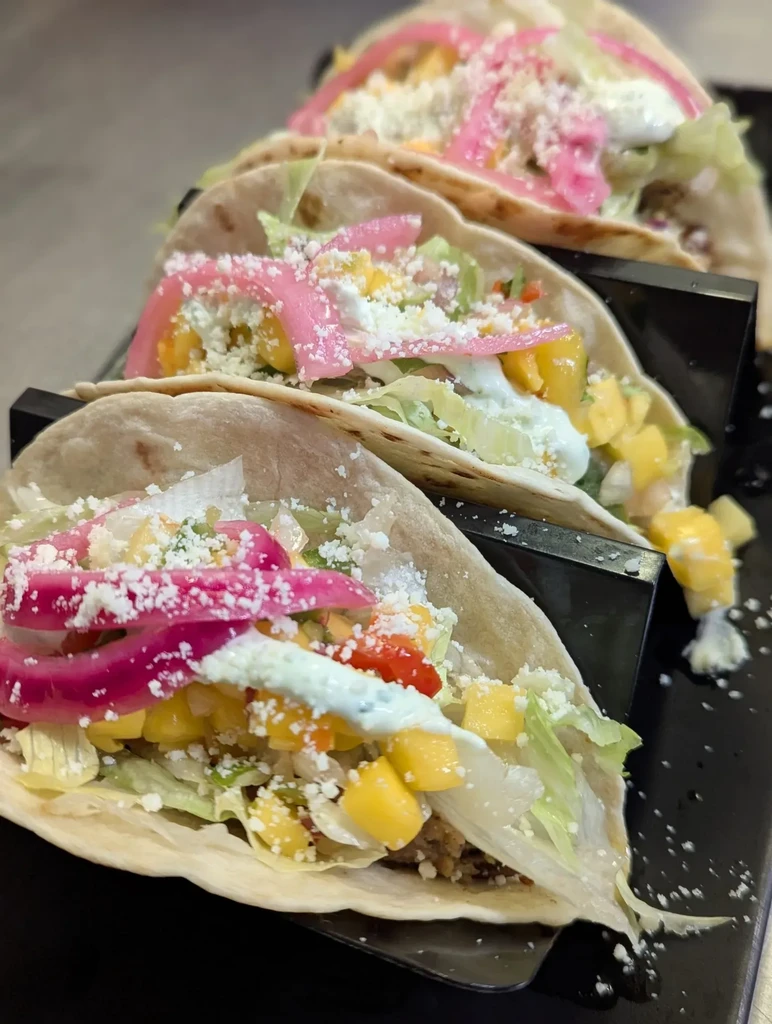 Three soft-shell tacos with mango, pickled onions, and cheese in a holder.