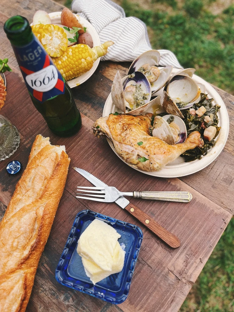 Rustic outdoor meal with roasted chicken, clams, corn, baguette, and 1664 beer.