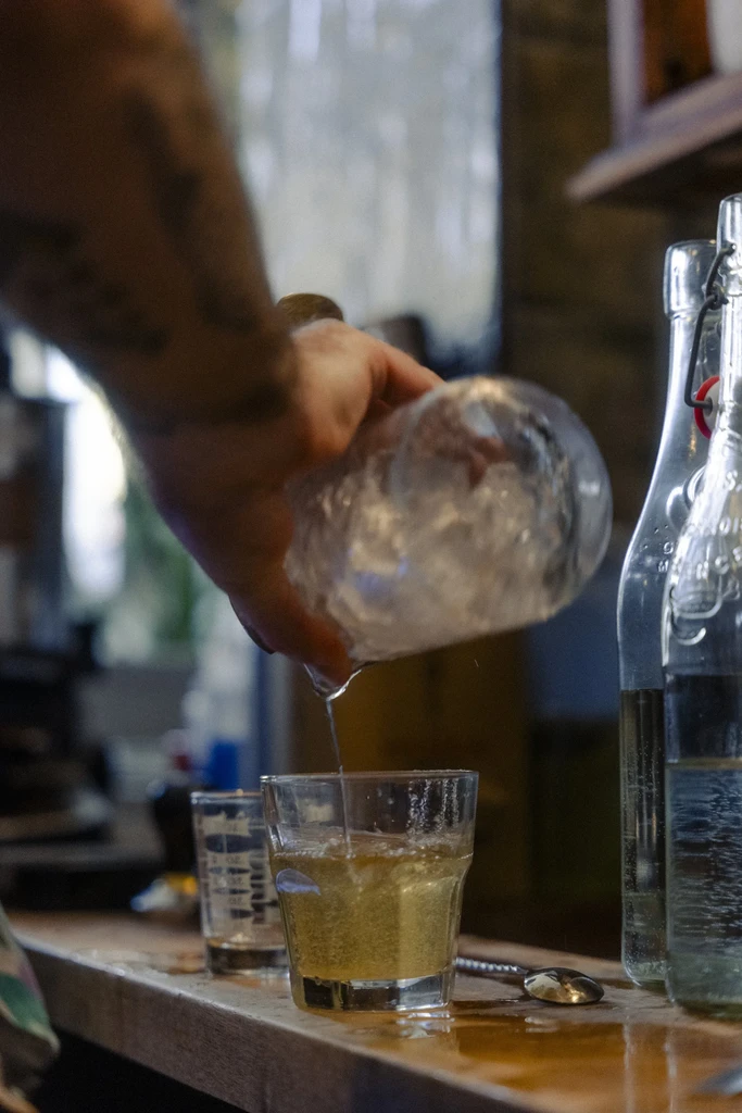 Tattooed hand pours liquid from carafe into a golden drink.
