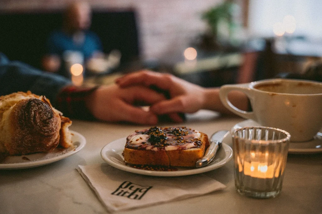 Cozy meal with jam toast, pastry, coffee, candle, and clasped hands.