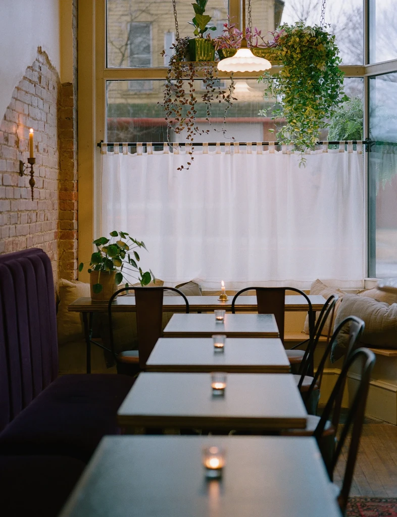 Restaurant interior: purple banquette, brick wall, candlelit tables, and potted plants.