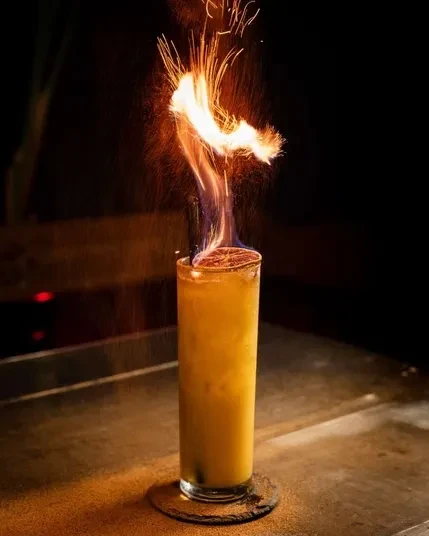 Flaming cocktail with charred citrus on a dark bar surface.