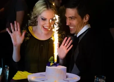 Two happy people look at a sparkling cake in a dark room.