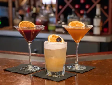 Three distinct cocktails, garnished with orange slices, on a bar.