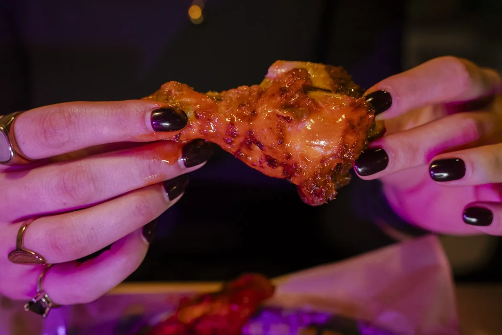 Hands with dark nails hold reddish-orange glazed chicken wing in purple light.