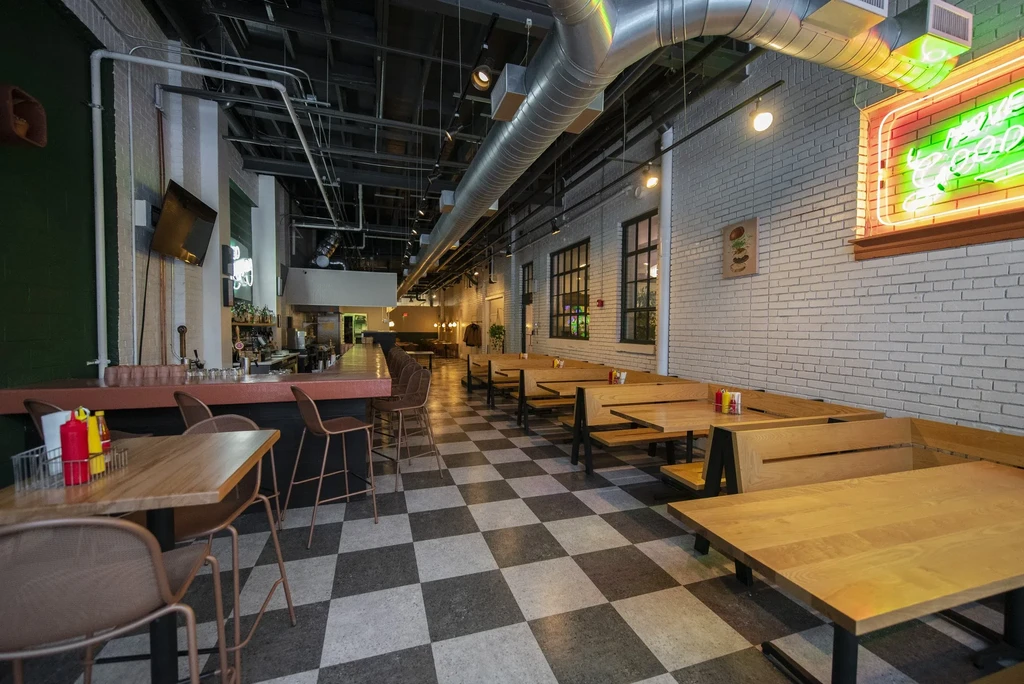 Industrial-chic restaurant interior with bar, booths, checkered floor, and neon sign.