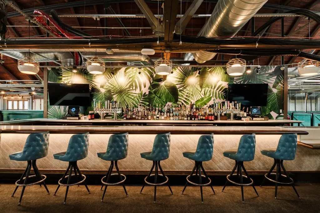 Modern bar with seven teal stools, tropical wallpaper, and industrial ceiling.
