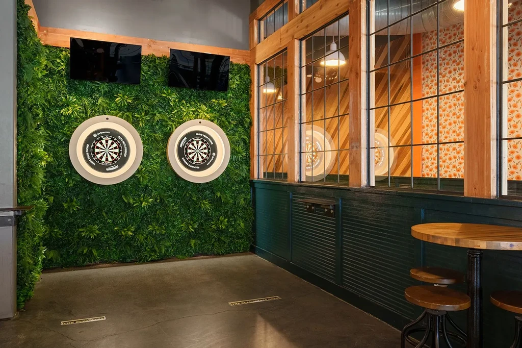 Two electronic dartboards on a green foliage wall in an entertainment space.