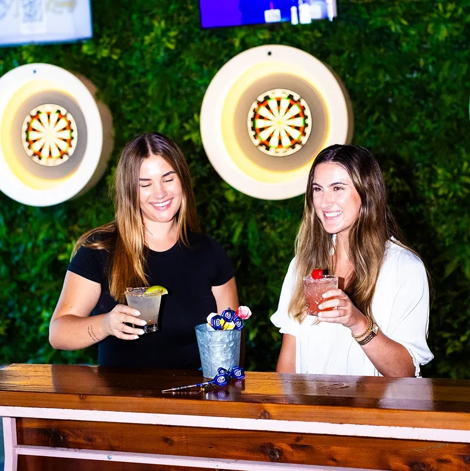 Smiling young women holding drinks at a bar with dartboards.