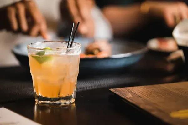 Close-up amber drink with ice, lime, straws; blurred dining background.