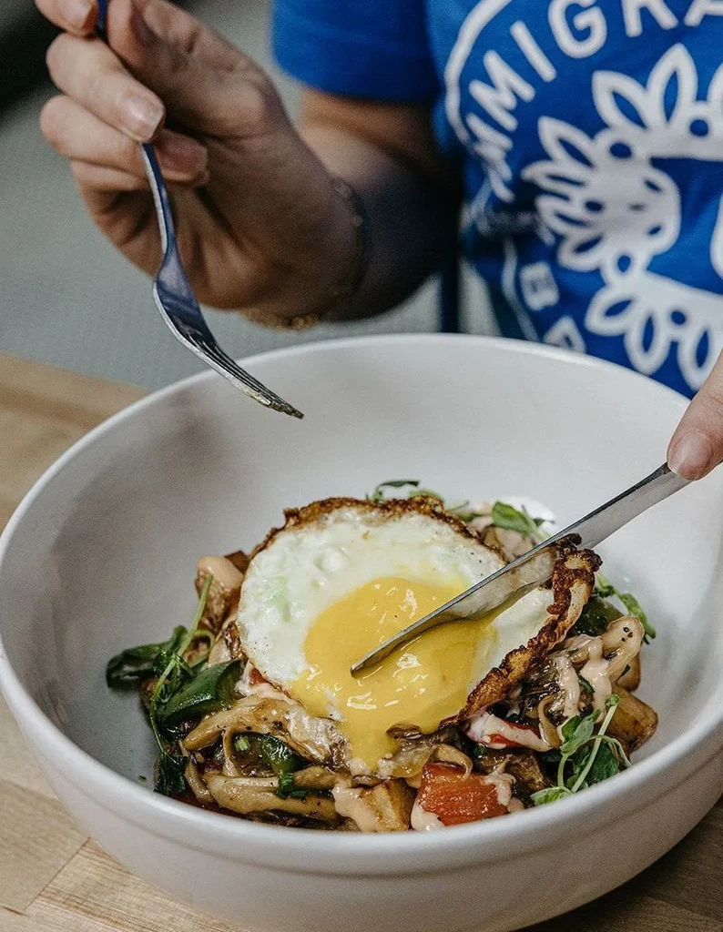 Knife cuts runny fried egg topping a hearty meal in a bowl.