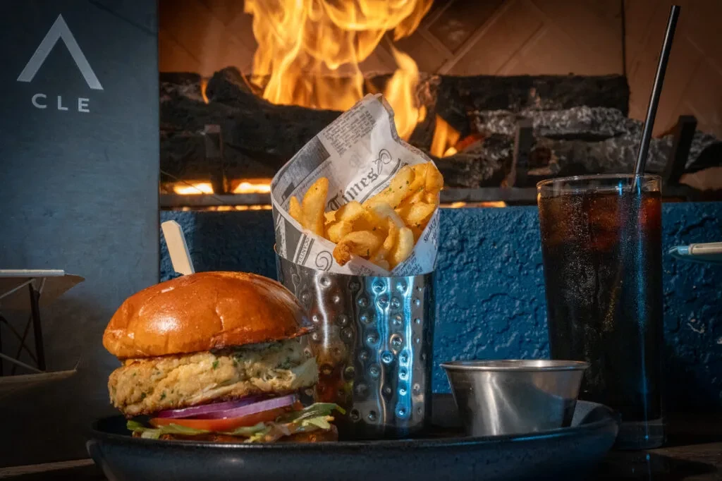 Close-up of a burger, curly fries, and dark drink by a fireplace.