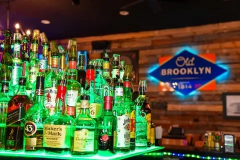 Green-lit liquor bottles at a bar, under an Old BROOKLYN neon sign.