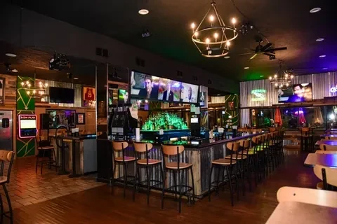 Modern bar with vibrant green bottle display, TVs, and industrial lights.