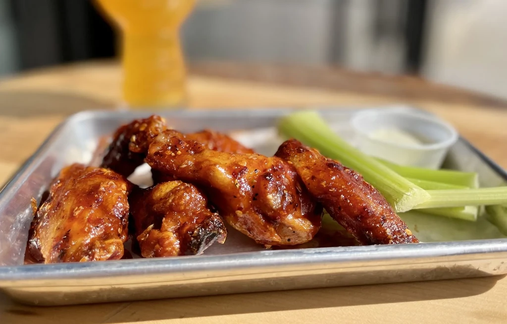 Glossy chicken wings, celery, and creamy dip on a metal tray.