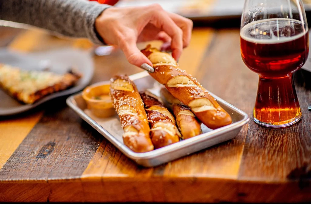 Hand takes a salted pretzel stick from tray with beer and dip.