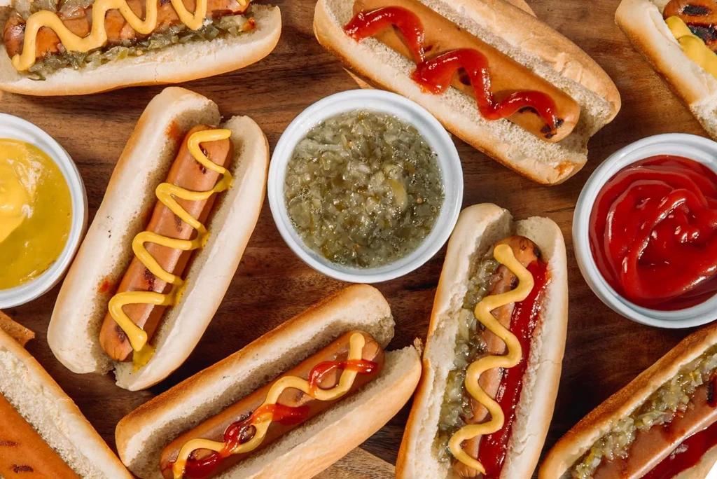 Overhead of hot dogs with mustard, ketchup, relish, and bowls on wood.
