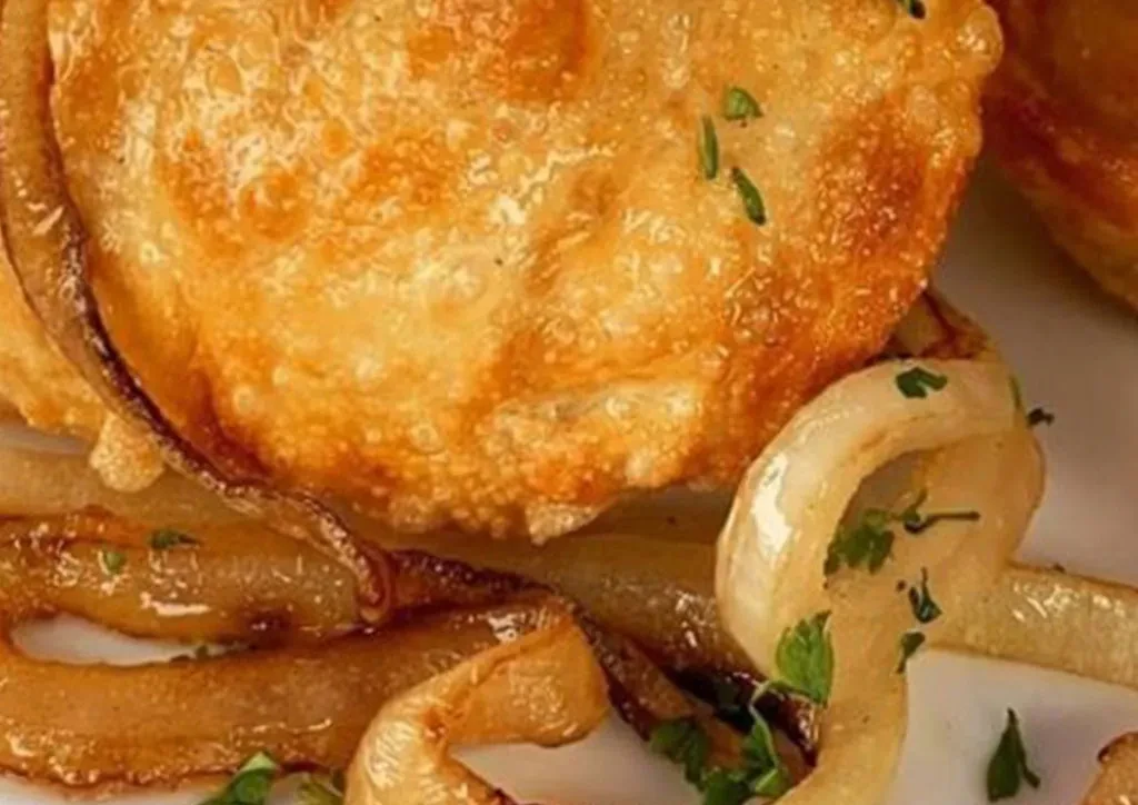 Close-up of golden-brown fried pastry with caramelized onions and green herbs.