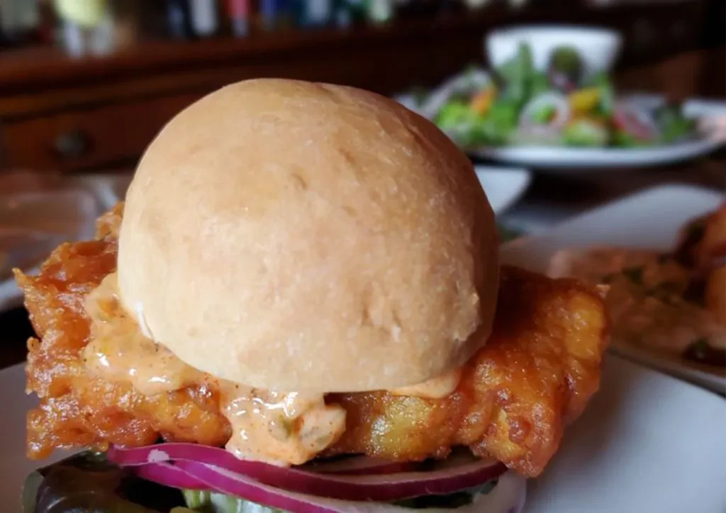 Close-up of a crispy fried fish sandwich with orange sauce and lettuce.