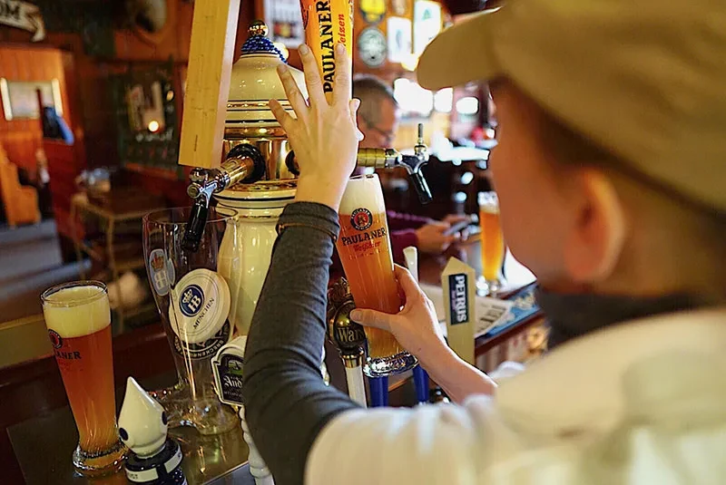 Person pouring Paulaner Weizen beer from a tap at a rustic pub.