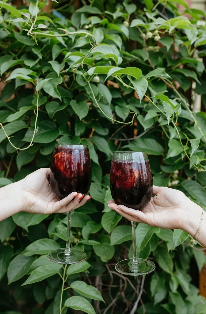 Two hands holding stemmed glasses with red drinks against green foliage.