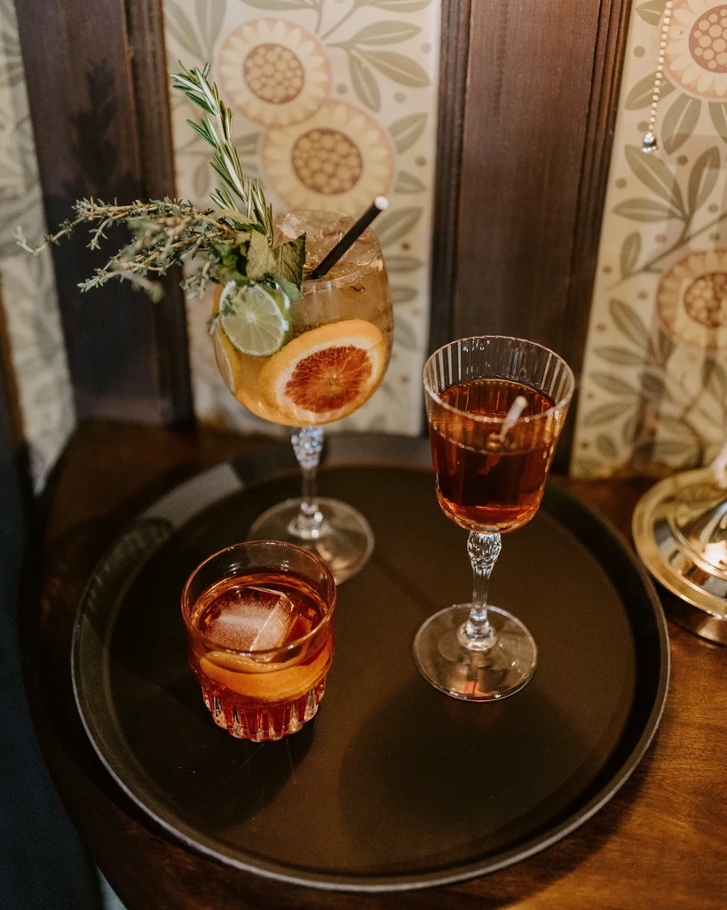 Three cocktails on a dark tray, with elaborate fruit and herb garnishes.