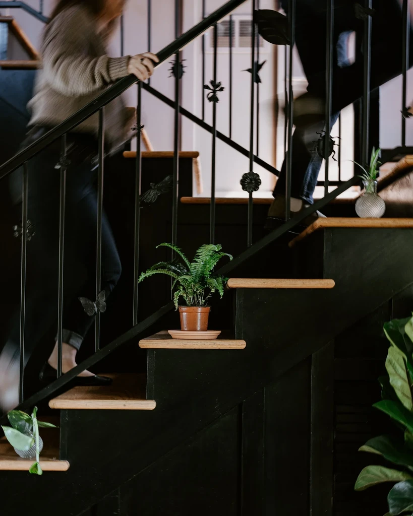 Dark staircase, iron railing with abstract motifs, plants, two blurred people.