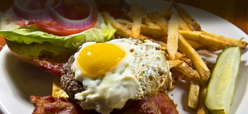 Open-faced burger with sunny-side egg, bacon, fries, and a pickle.