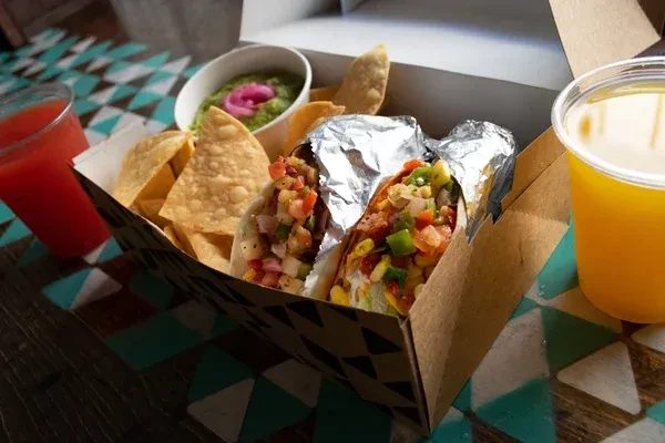 Takeaway box with two colorful tacos, chips, guacamole, and two drinks.