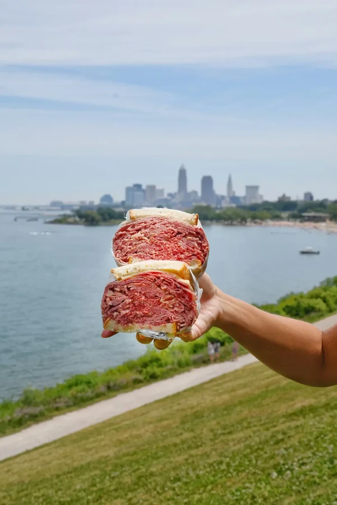 Hand holds large deli sandwich with city skyline over water.