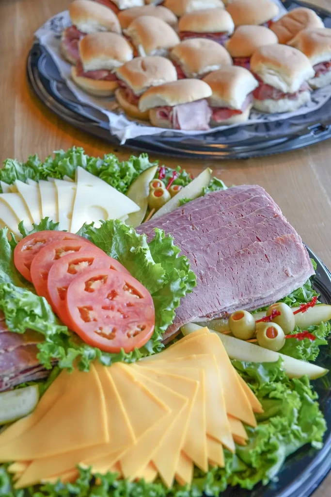 Two black trays: deli platter with meat, cheese, and slider sandwiches.