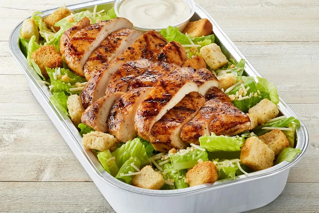 Grilled chicken Caesar salad in a takeout container on wood.