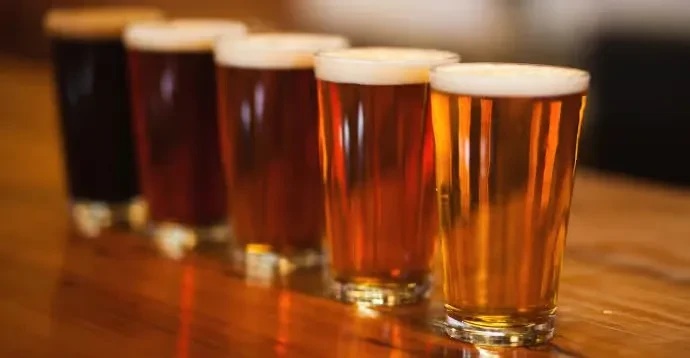 Six pint glasses of beer, dark to light, on a wooden bar.