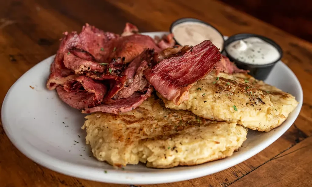 Sliced meat, golden potato patties, and two sauces on a white plate.