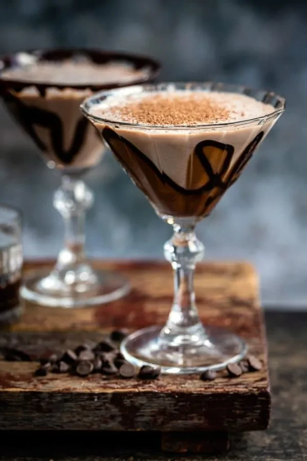 Creamy chocolate martini with dark syrup, shavings, and chips on wood.