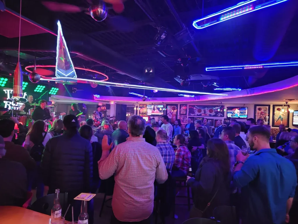 Crowded bar scene with a band on stage and vibrant neon lights.