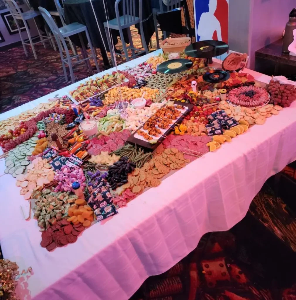 Large, colorful grazing table under pink light, laden with diverse foods.